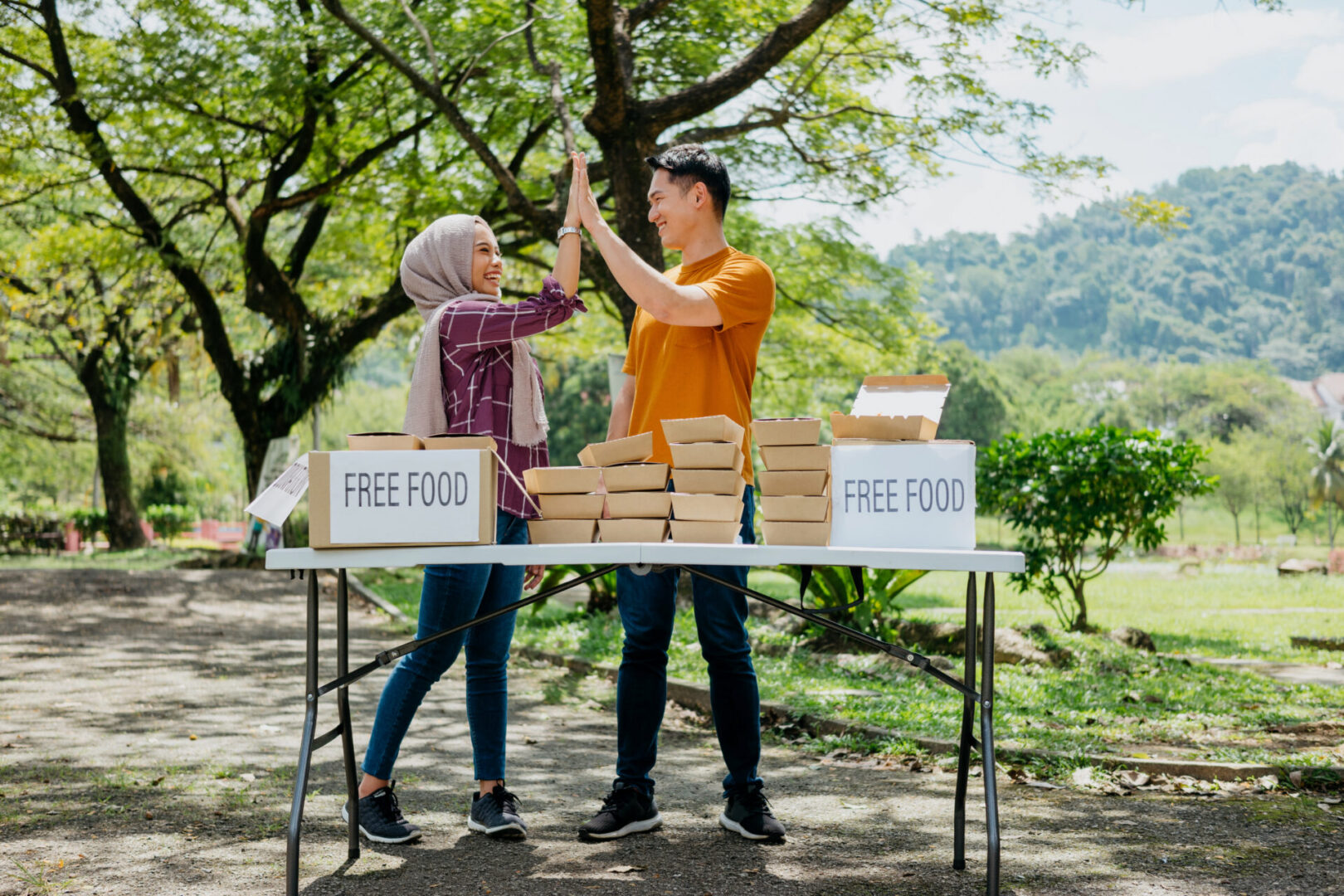 Image of two young asian volunteers ready to give free food to those impacted by financial difficulty at public park. They are giving each other a high five for completing the preparation.