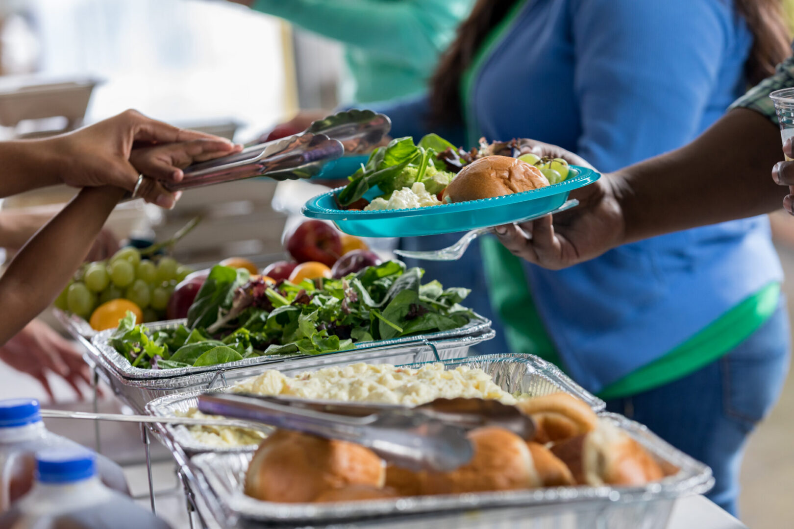 Volunteer serves healthy meal to an unrecognizable man in soup kitchen. The volunteer is placing green salad, potato salad and a roll on his plate. Close up is on the serving tongs and plate.
